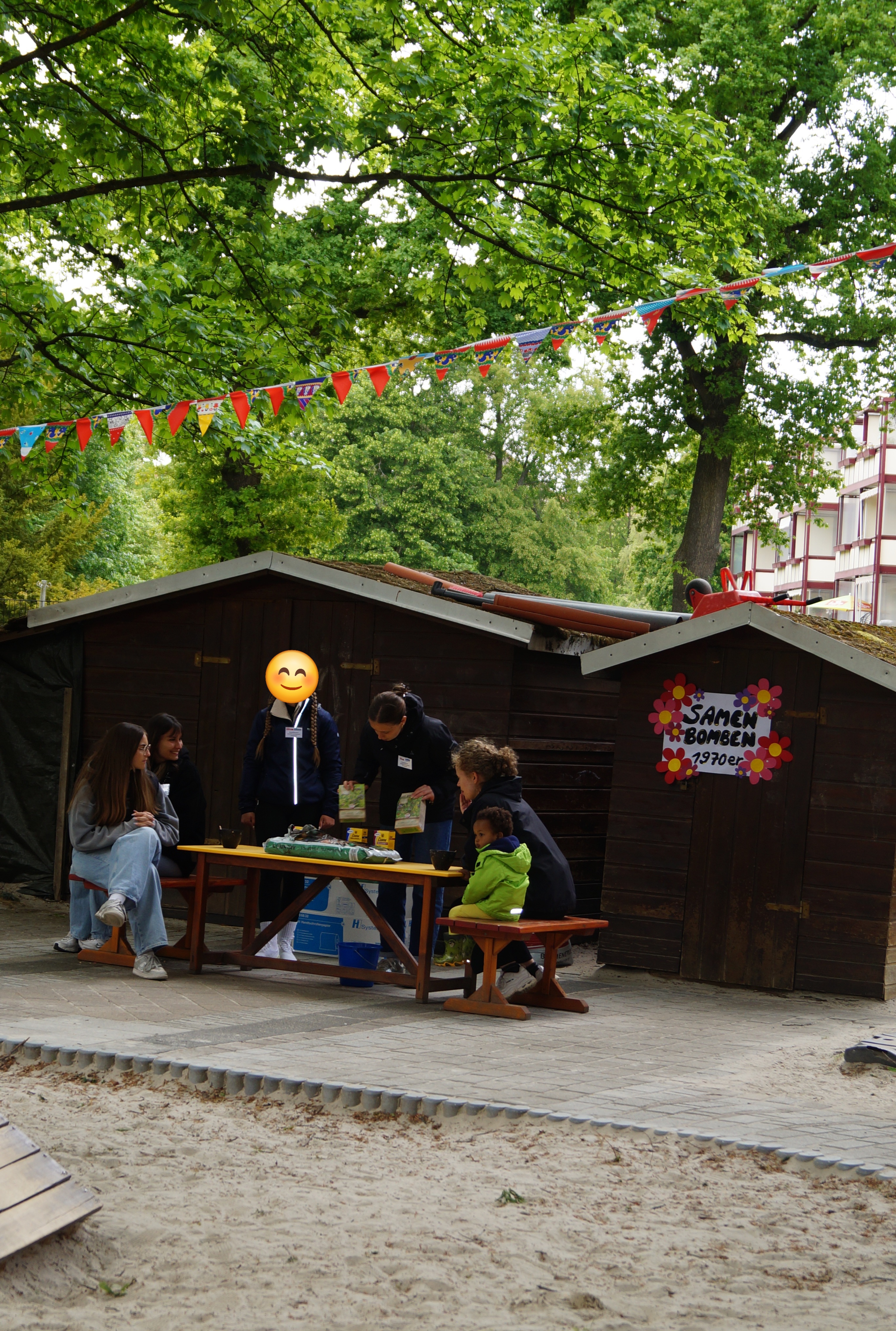 Ein Außengelände mit einem Holzhaus, an dem bunte Wimpel hängen. Mehrere Erwachsene und ein Kind sitzen an einem Tisch und unterhalten sich. Am Haus ist ein bunt gestaltetes Schild mit der Aufschrift "Samenbomben" angebracht, was auf eine Aktivität oder einen Stand hindeutet.