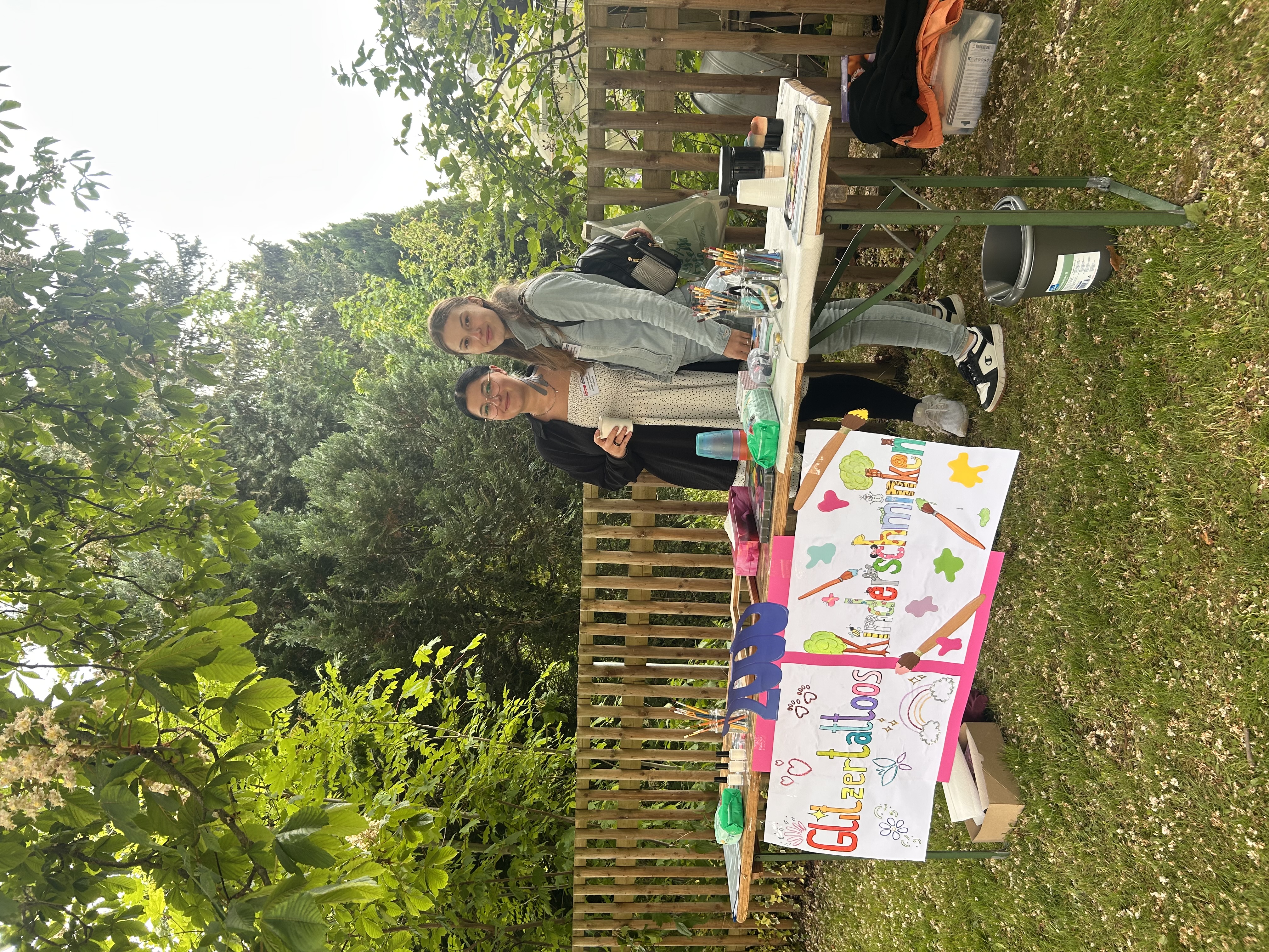 Zwei Personen stehen freundlich lächelnd hinter einem Tisch im Freien, auf dem ein bunt gestaltetes Schild steht, das auf Glitzertattoos und Kinderschminken hinweist. Der Tisch ist ausgestattet mit Farben, Pinseln und Zubehör für die Aktivitäten. Die Umgebung ist von viel Grün umgeben und wirkt freundlich und einladend.