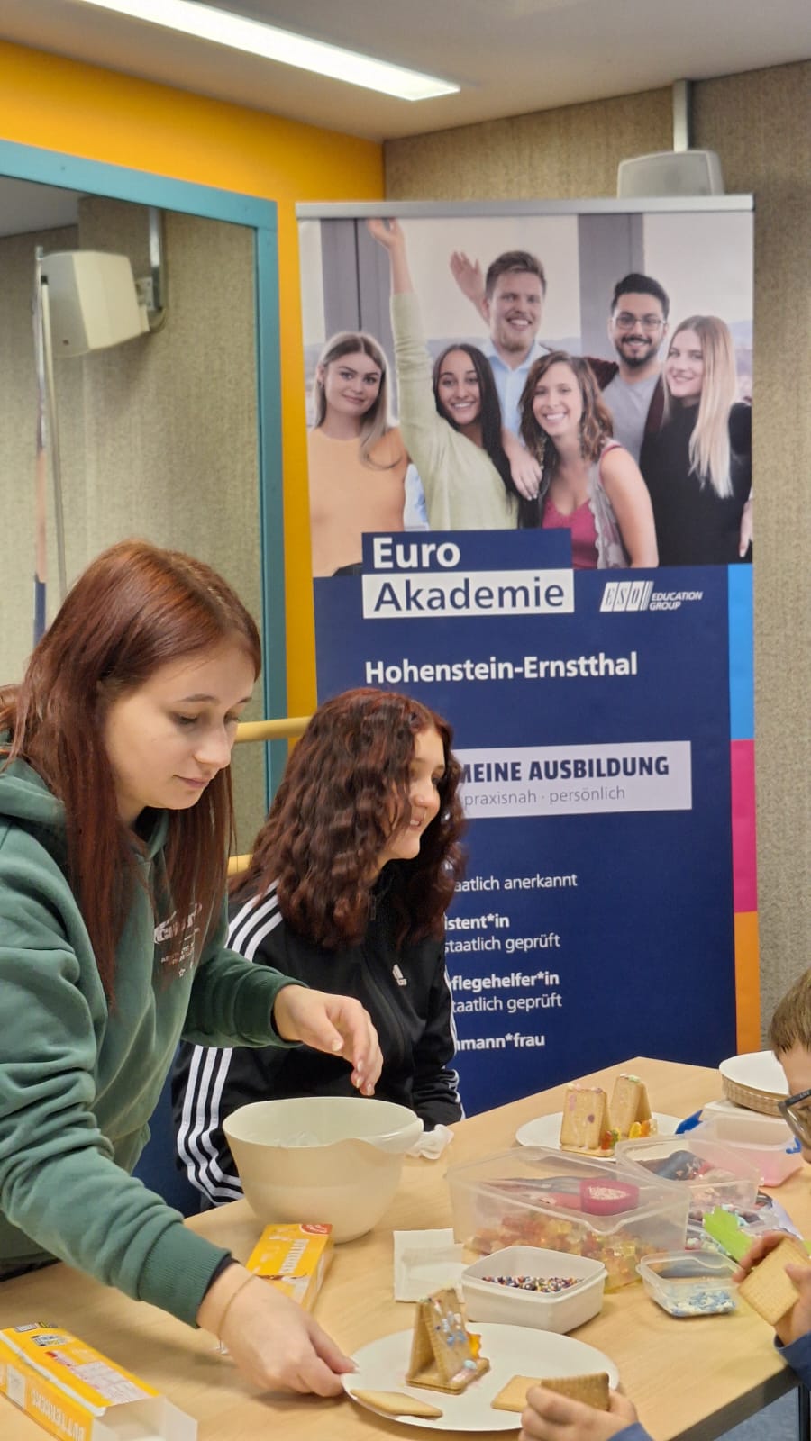 Zwei junge Frauen sitzen mit Kindern an einem Tisch und bauen kleine Kekshäuschen; vor ihnen stehen Schüsseln, Teller und Bastelmaterial, im Hintergrund ist ein großes Roll-up der Euro Akademie Hohenstein-Ernstthal mit lächelnden jungen Menschen zu sehen.