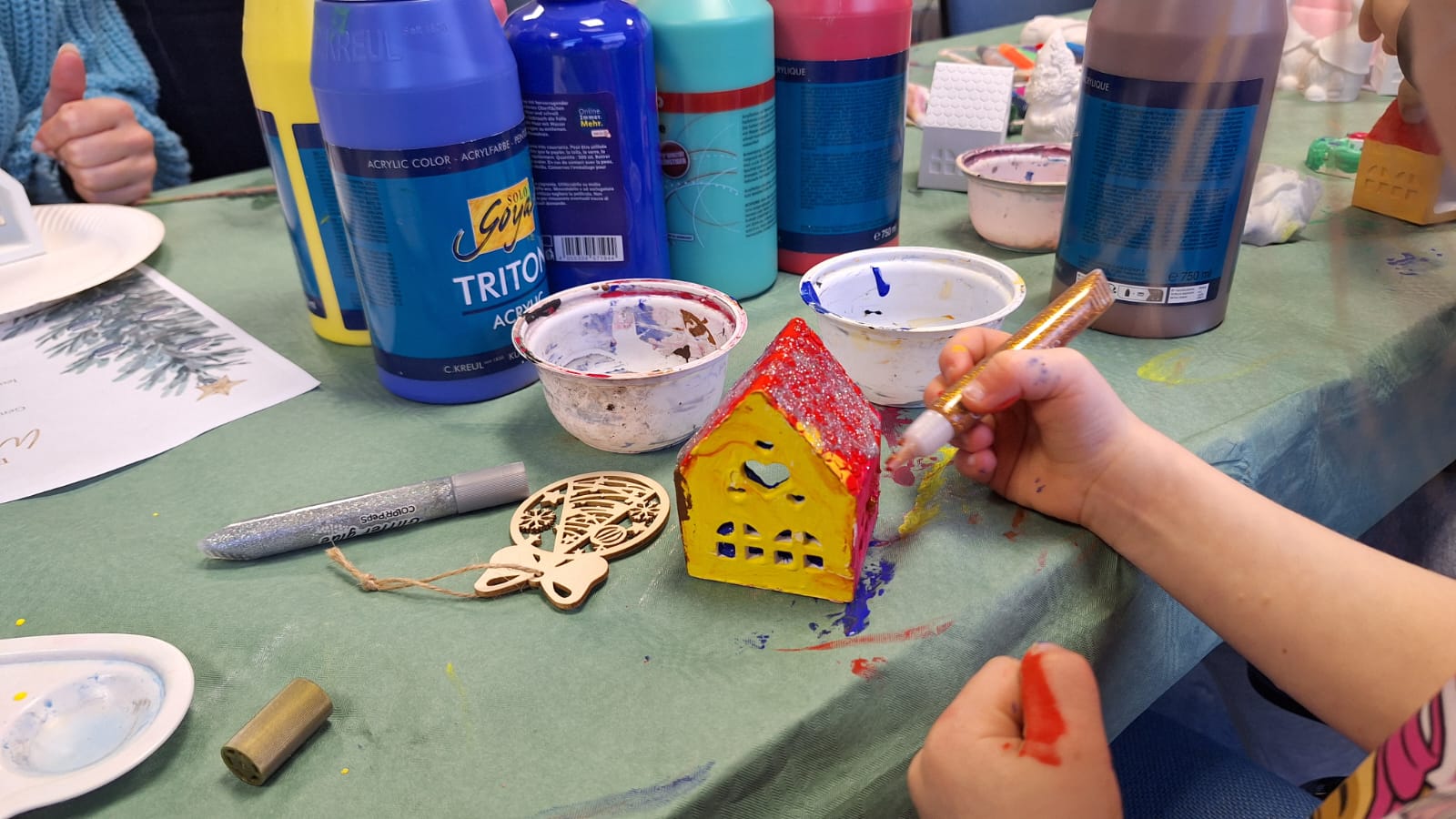 Ein Kind bemalt ein kleines Keramikhäuschen in Rot und Gelb mit einem goldenen Stift, auf dem Tisch stehen mehrere große Flaschen Acrylfarbe und Schalen mit Farbresten sowie weihnachtliche Holzanhänger.