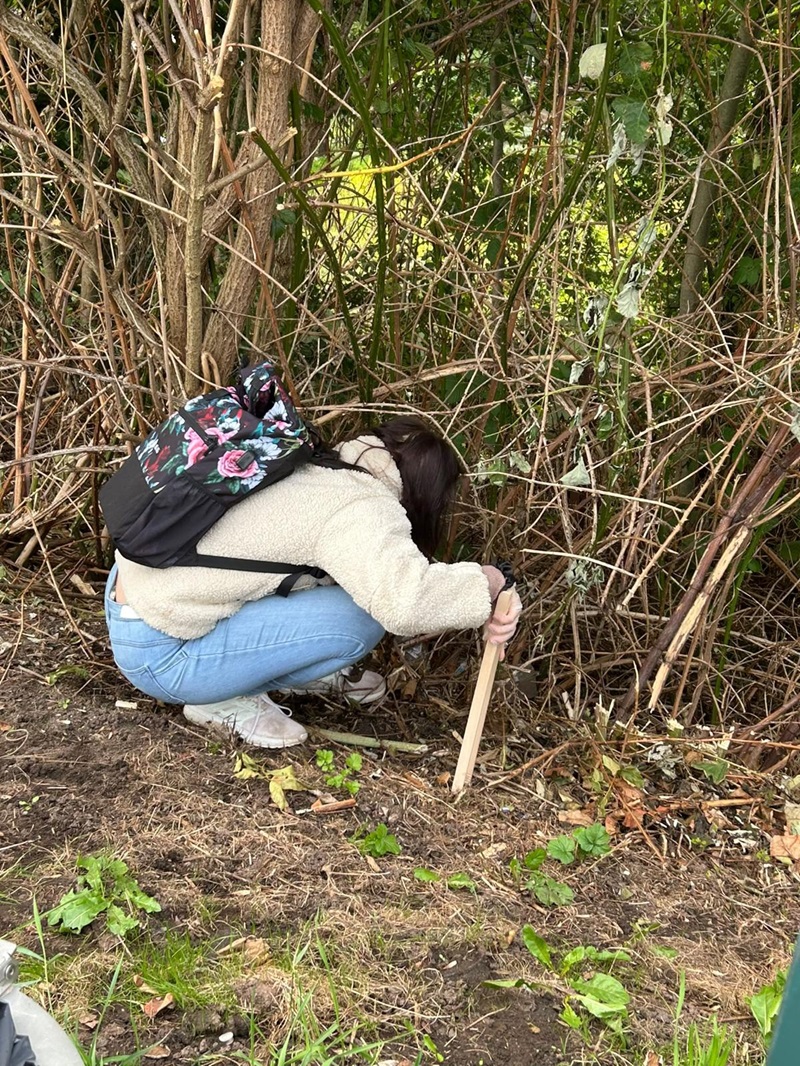 Eine Schülerin mit einem cremefarbenen Pullover, hellblauer Jeans und weißen Schuhen hockt am Rand eines Gebüschs. Sie trägt einen schwarzen Rucksack mit Blumenmuster und hält einen Holzstab in der Hand, den sie in den Boden steckt. Im Hintergrund sind dichte, braune und grüne Äste und Blätter zu sehen. Der Boden ist teilweise mit Gras und Laub bedeckt.