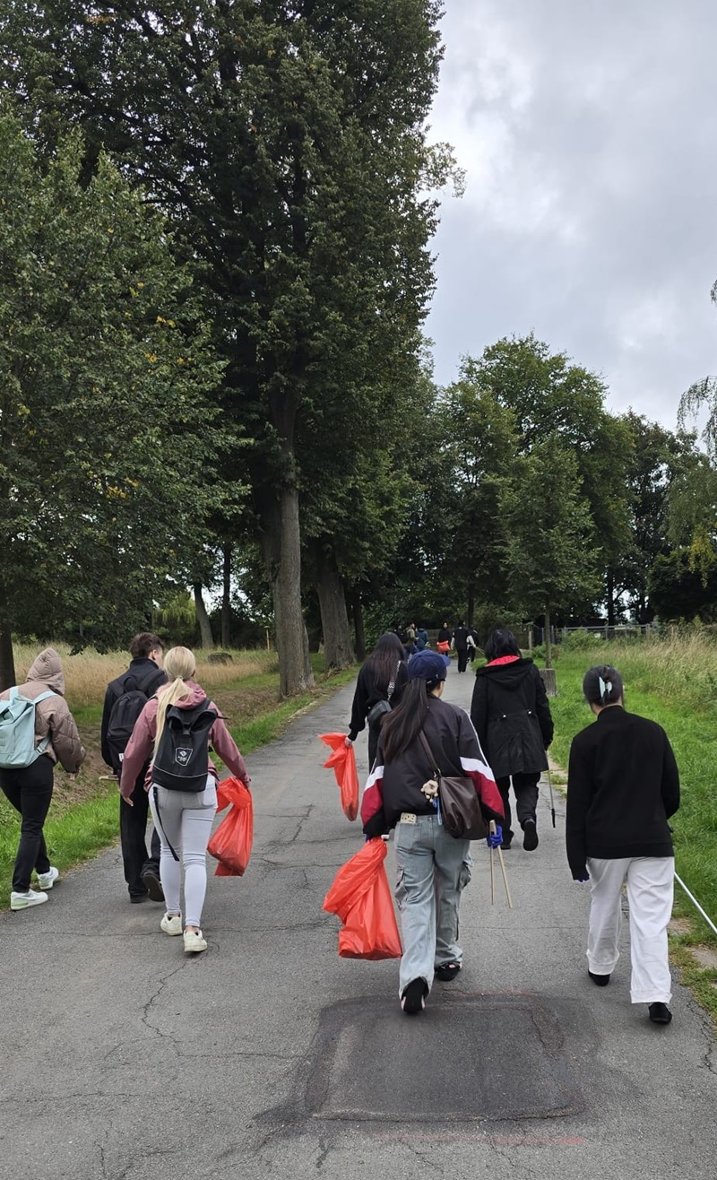 Eine Gruppe von Schülerinnen und Schülern geht auf einem asphaltierten Weg durch eine grüne Parklandschaft. Einige tragen orangefarbene Müllsäcke und sind mit Rucksäcken ausgestattet. Die Schülerinnen und Schüler sind von hinten zu sehen und tragen überwiegend sportliche oder legere Kleidung. Links und rechts des Weges stehen hohe Bäume, der Himmel ist bewölkt.