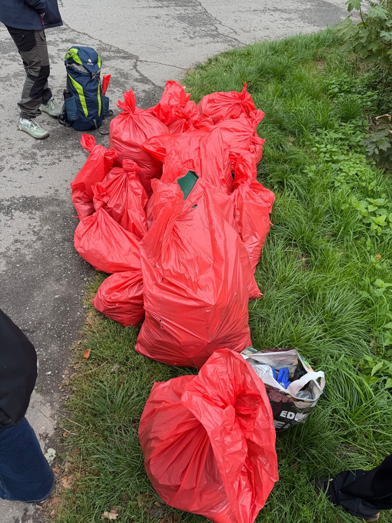 Mehrere rote Müllsäcke liegen auf einer Grasfläche am Rand einer asphaltierten Straße. Ein Wanderrucksack steht neben einer Person, deren Beine sichtbar sind. Im Vordergrund befindet sich eine offene Einkaufstasche mit verschiedenen Gegenständen. Die Szene vermittelt den Eindruck einer Sammelaktion im Freien.