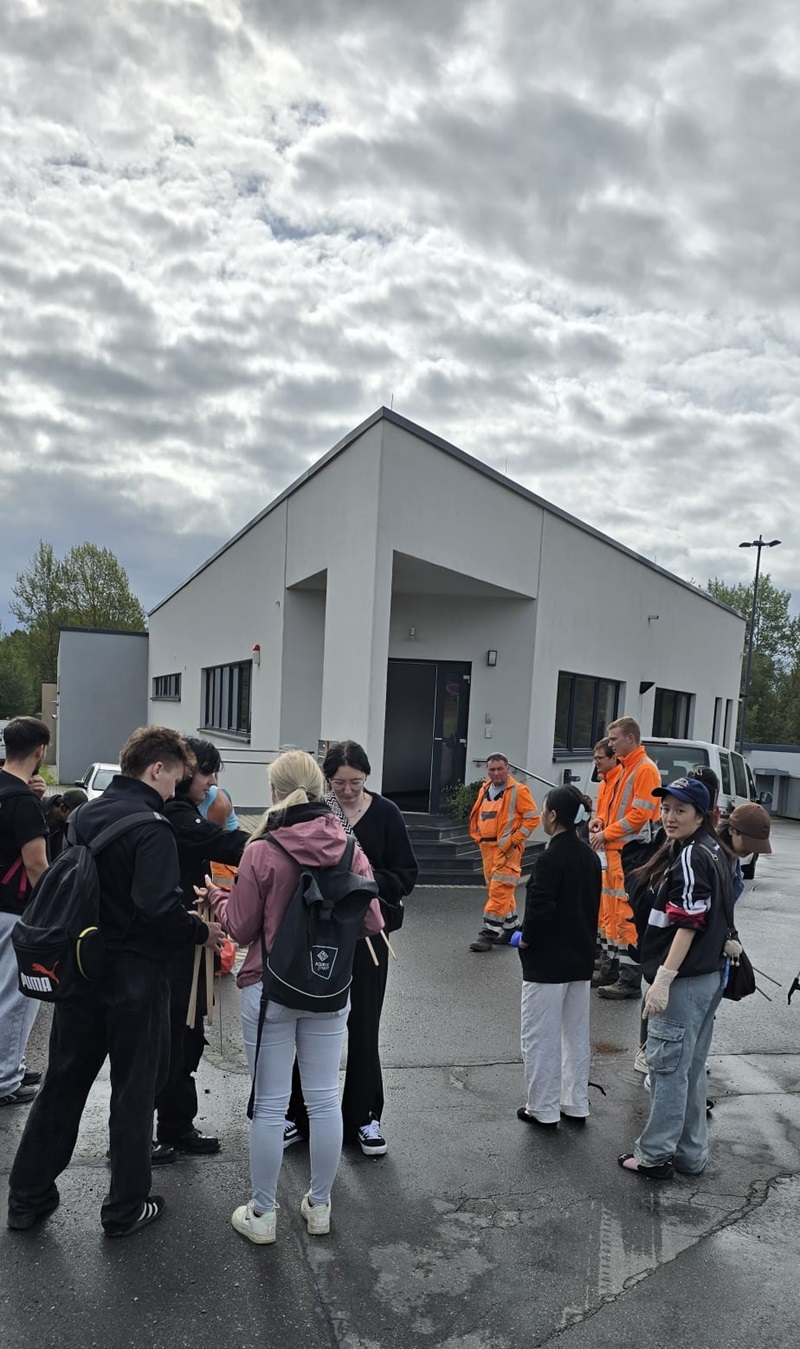Vor einem modernen, weiß gestrichenen Gebäude mit spitzem Dach stehen mehrere Schülerinnen und Schüler auf einem nassen Asphaltplatz. Einige tragen dunkle Freizeitkleidung und Rucksäcke, andere sind in auffälligen orangefarbenen Arbeitsanzügen gekleidet. Der Himmel ist von dichten Wolken bedeckt, wodurch das Licht diffus wirkt. Im Hintergrund sind Bäume und weitere Gebäudeteile zu erkennen.