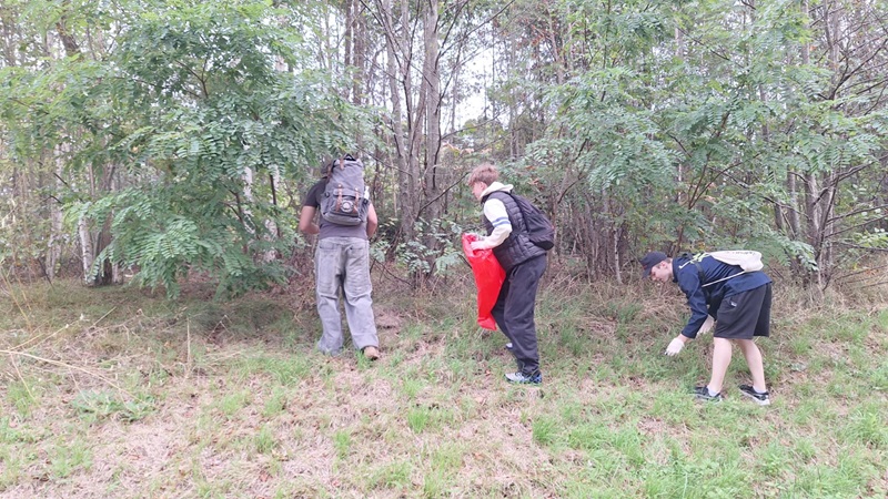 Drei Schüler stehen oder hocken auf einer Wiese am Waldrand. Sie tragen Rucksäcke und Outdoor-Kleidung. Eine Person hält einen roten Stoff oder Beutel, die andere bückt sich und sammelt etwas vom Boden auf. Im Hintergrund sind dichte Bäume und Sträucher zu sehen.