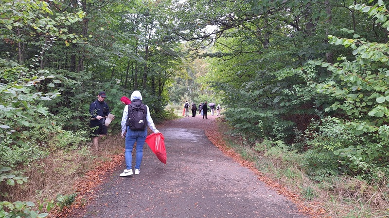Mehrere Schüler gehen auf einem asphaltierten Weg durch einen dichten, grünen Laubwald. Einige tragen Rucksäcke und wetterfeste Kleidung, eine Person hält einen großen roten Müllsack. Der Boden ist feucht und mit herabgefallenem Laub bedeckt. Die Szene wirkt herbstlich und ruhig.