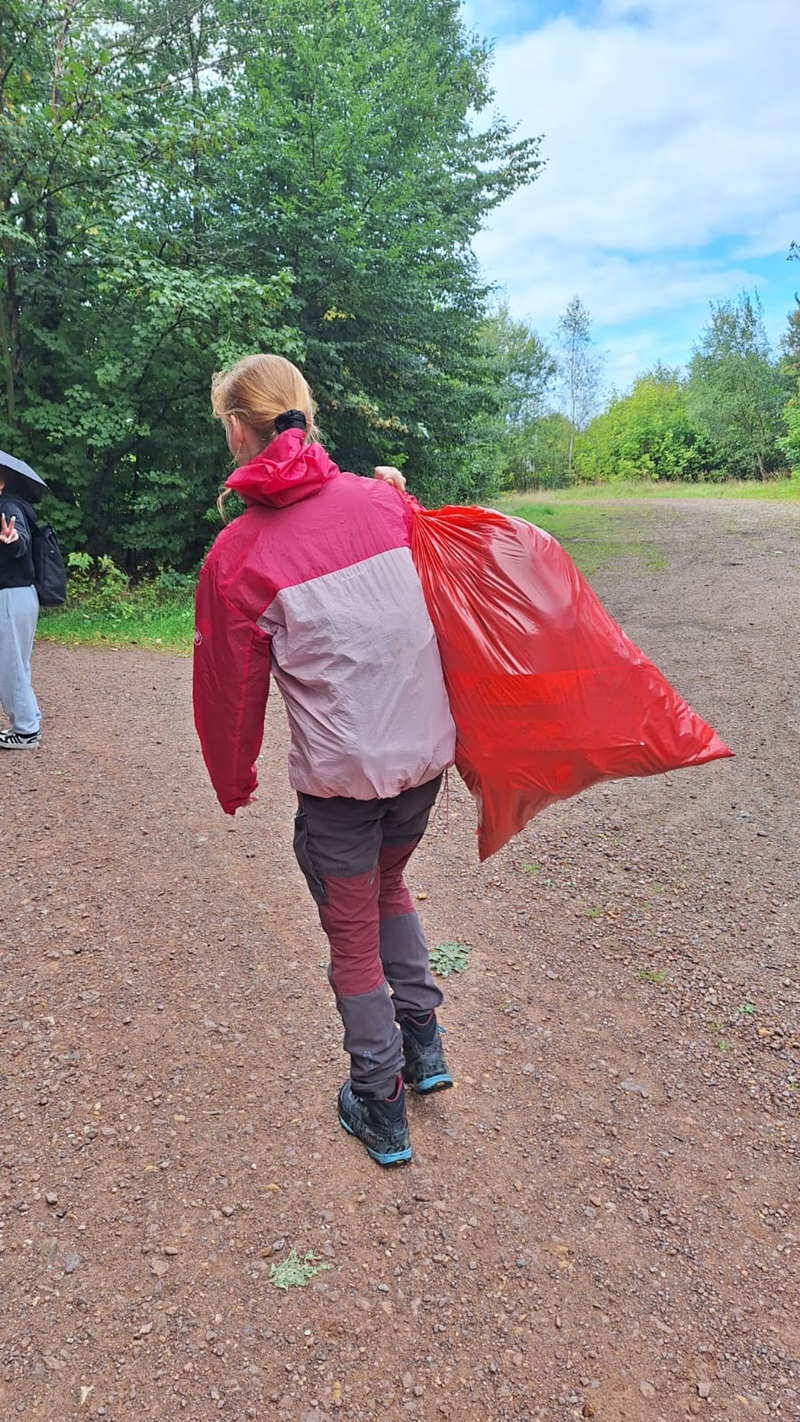 Eine Schülerin mit blondem Haar trägt eine rot-rosa Outdoorjacke und eine dunkle Hose. Sie hält einen großen, leuchtend roten Müllsack über der Schulter. Die Szene spielt auf einem unbefestigten Weg, umgeben von grünen Bäumen. Im Hintergrund steht eine weitere Person mit dunkler Kleidung und einem Regenschirm.