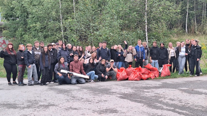 Eine große Gruppe junger Schülerinnen und Schüler steht auf einem asphaltierten Platz vor einer Baumreihe. Die Personen tragen überwiegend dunkle Freizeitkleidung und lächeln oder winken in Richtung Kamera. Vor der Gruppe liegen mehrere rote Müllsäcke, die gefüllt wirken. Einige Personen halten längliche Gegenstände in den Händen. Die Szene vermittelt den Eindruck einer gemeinschaftlichen Aktivität im Freien.