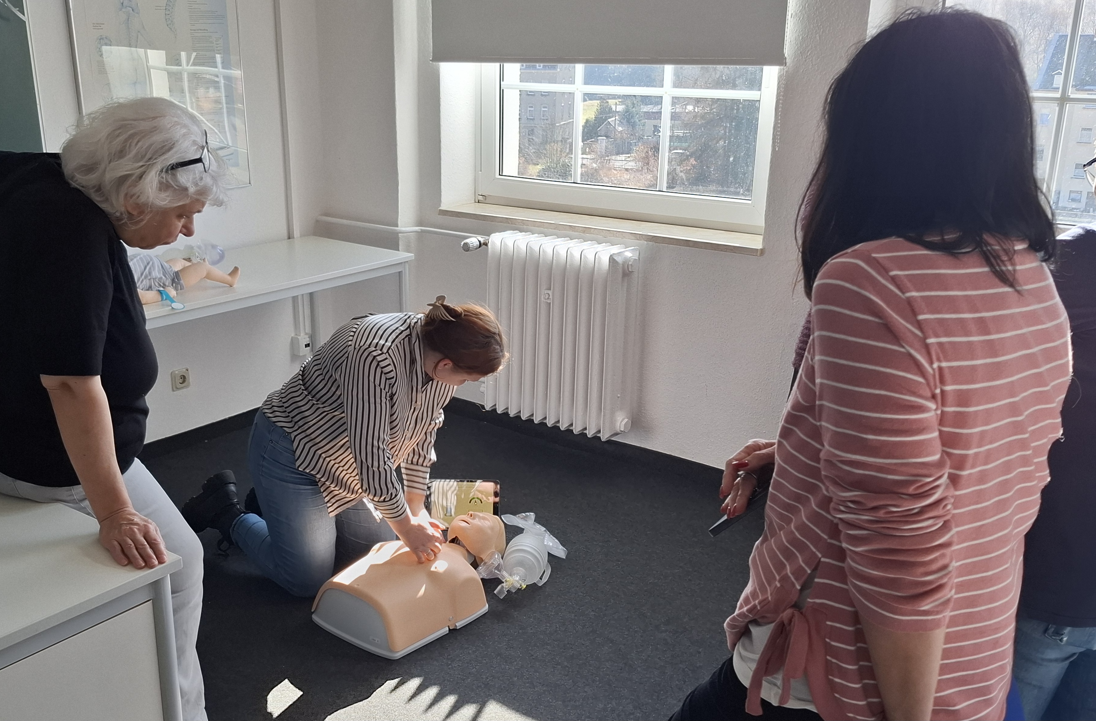 Mehrere Erwachsene nehmen in einem hellen Unterrichtsraum an einer Erste-Hilfe-Übung teil, während eine Teilnehmerin an einer Reanimationspuppe die Herzdruckmassage demonstriert.