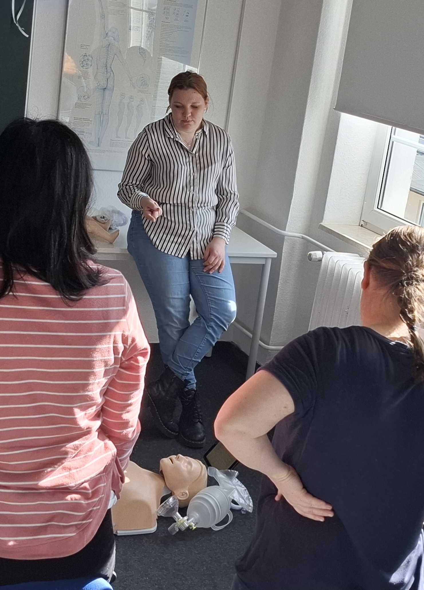 Eine Teilnehmerin erklärt in einem Schulungsraum die Wiederbelebung mit einer Reanimationspuppe, während andere Personen aufmerksam zuschauen.