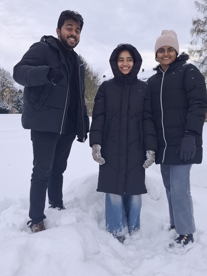 Die Auszubildenden stehen im Schnee. Alle tragen dunkle Winterjacken, Handschuhe und Mützen. Die mittlere Person trägt eine lange, schwarze Daunenjacke und helle Jeans, die am Saum vom Schnee bedeckt sind. Die rechte Person trägt eine helle Strickmütze, eine schwarze Jacke und hellblaue Hosen. Die linke Person trägt eine schwarze Jacke, schwarze Hose und hebt die rechte Hand leicht an. Im Hintergrund sind schneebedeckte Bäume und eine offene Schneefläche zu sehen.