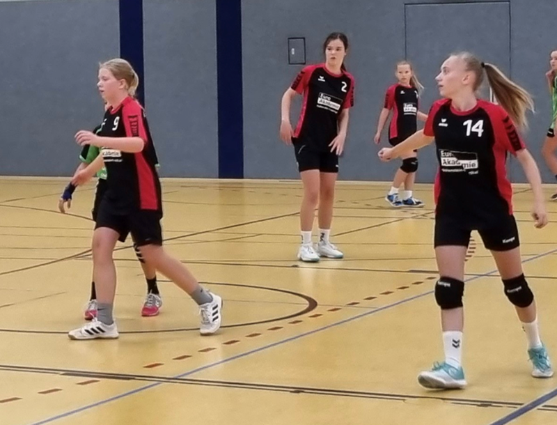 Mehrere Handballspielerinnen in schwarzen Trikots mit roten Streifen stehen auf einem Hallenboden mit markierten Spielfeldlinien. Die Trikots tragen weiße Nummern und einen weißen Schriftzug auf der Brust. Eine Person trägt eine schwarze Kniebandage und weiße Sportschuhe. Im Hintergrund sind blaue und graue Wandflächen zu sehen.