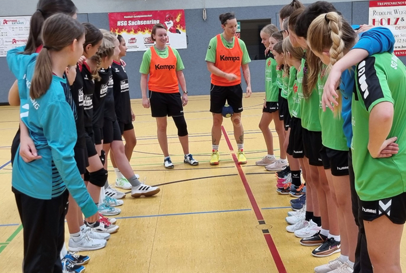 Eine Gruppe junger Handballspieler steht sich in einer Sporthalle gegenüber. Links hört ein Team in blau-schwarzen Trikots mit weißen Schuhen, einige tragen Knieschoner, aufmerksam zu. Rechts steht ein Team in grünen Trikots mit bedruckten schwarzen Shorts und verschiedenen Schuhen, einige mit Pferdeschwänzen, eng beieinander. In der Mitte unterhalten sich zwei Erwachsene in grünen Hemden und orangefarbenen Westen mit den Teams. Es scheint sich um ein Training zu handeln, Banner sind zu sehen.