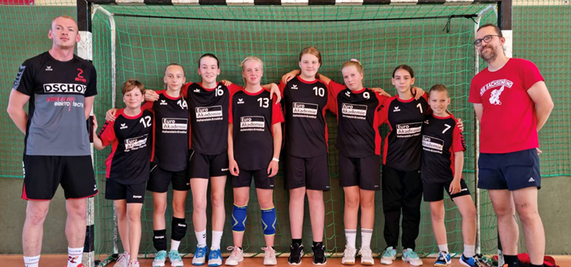 Eine Gruppe junger Handballspieler steht vor einem Tor in einer Turnhalle. Sie tragen einheitliche schwarz-rote Sportuniformen mit gut sichtbaren Nummern und Sponsorenlogos. Zwei Erwachsene, vermutlich Trainer, stehen jeweils links und rechts von ihnen. Der Trainer links trägt eine andere Uniform, der Trainer rechts ein rotes T-Shirt und Shorts. Das Team posiert lächelnd für ein Mannschaftsfoto.