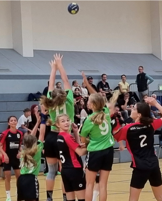 In einer Sporthalle findet gerade ein Handballspiel statt. Spielerinnen in grünen Trikots mit den Nummern 7 und 9 springen nach dem Ball, während auch Spieler in schwarzen und roten Trikots im Einsatz sind. Zuschauer sitzen im Hintergrund und verfolgen das Spiel gespannt. Die Szene fängt die Energie und den Kampfgeist des Spiels perfekt ein.