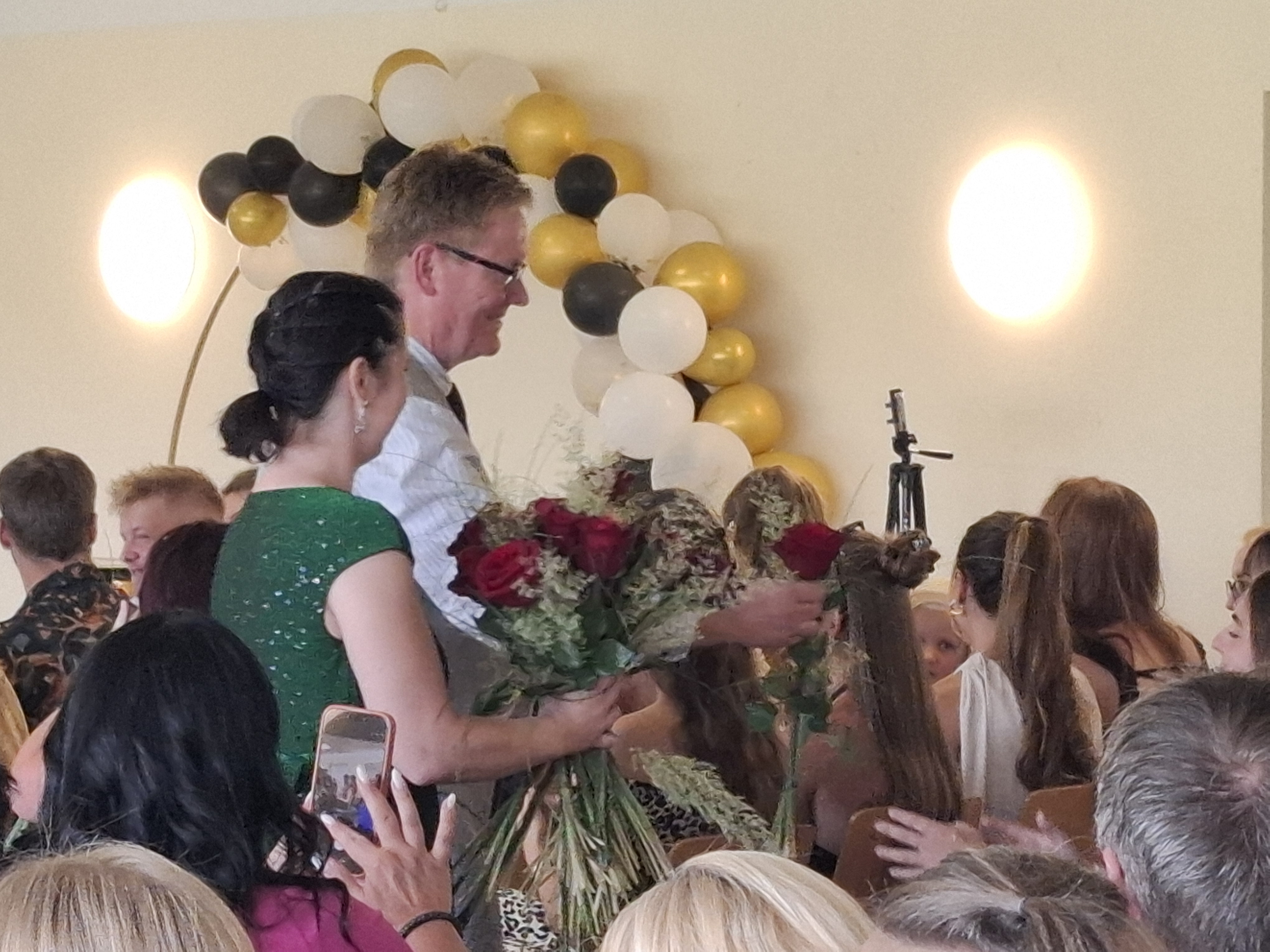 Eine Frau und ein Mann mit Blumensträußen in den Händen stehen in einer Zuschauermenge. Im Hintergrund hängt eine Girlande aus schwarzen, weißen und goldenen Luftballons.