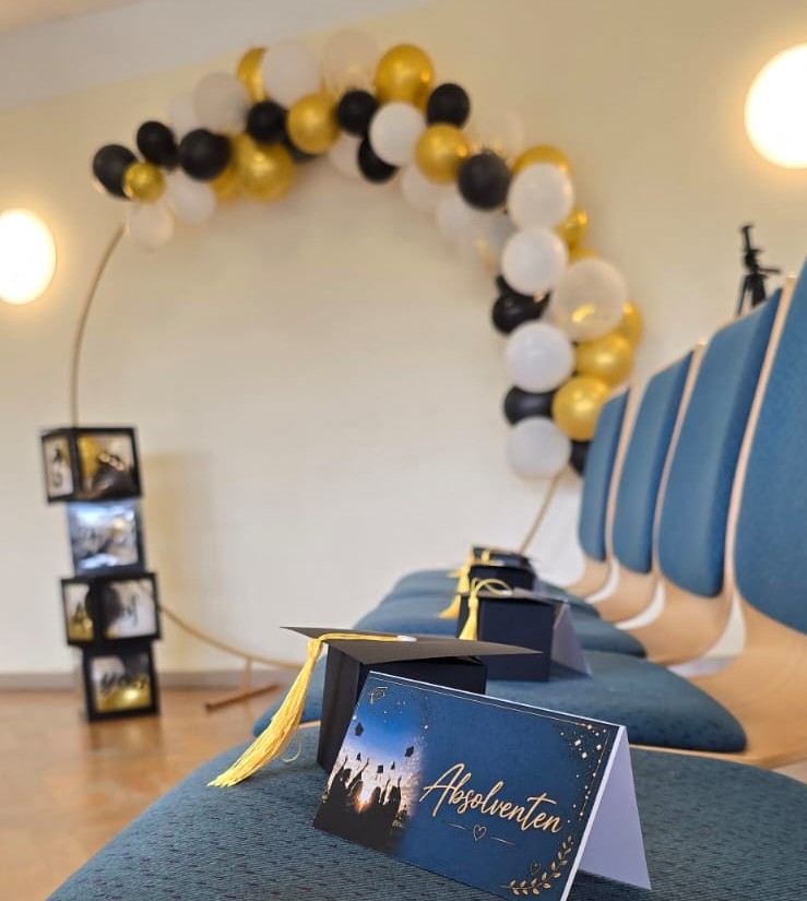 Auf einer Reihe blauer Stühle liegen schwarze Absolventenhüte, ganz vorne steht eine blau-goldene Klappkarte mit der Aufschrift "Absolventen". Im Hintergrund hängt eine Girlande aus schwarzen, weißen und goldenen Luftballons.