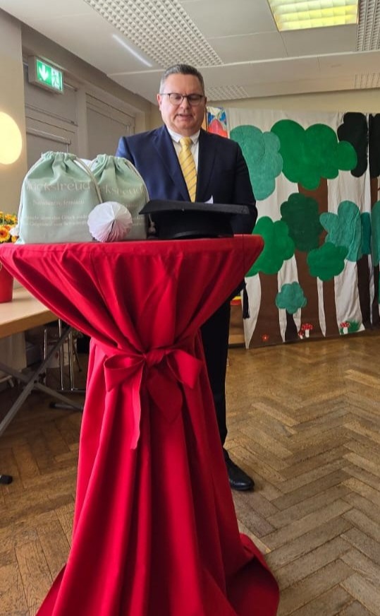 An einem Stehtisch mit einem roten Überzug, auf dem ein Absolventenhut liegt, steht ein Mann in Anzug und gelber Krawatte. Im Hintergrund ist eine mit Bäumen bemalte Wand.