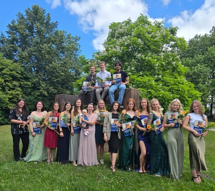 Auf einer grünen Wiese mit Bäumen im Hintergrund stehen junge Menschen in Abendkleidern für ein Gruppenbild; drei davon sitzen erhöht auf einem Stein. Alle halten jeweils eine Blume und ein Zeugnis in der Hand.