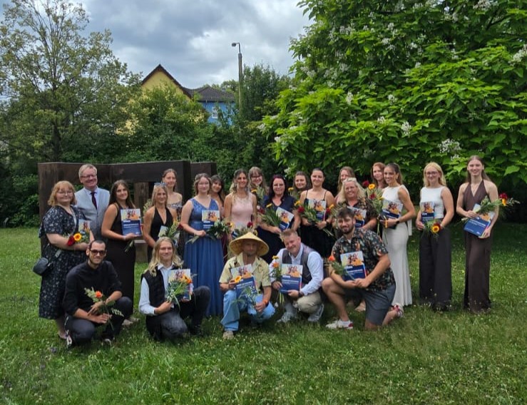 Auf einer grünen Wiese mit Bäumen im Hintergrund stehen junge Menschen in Abendkleidern für ein Gruppenbild. Alle halten jeweils eine Blume und ein Zeugnis in der Hand.