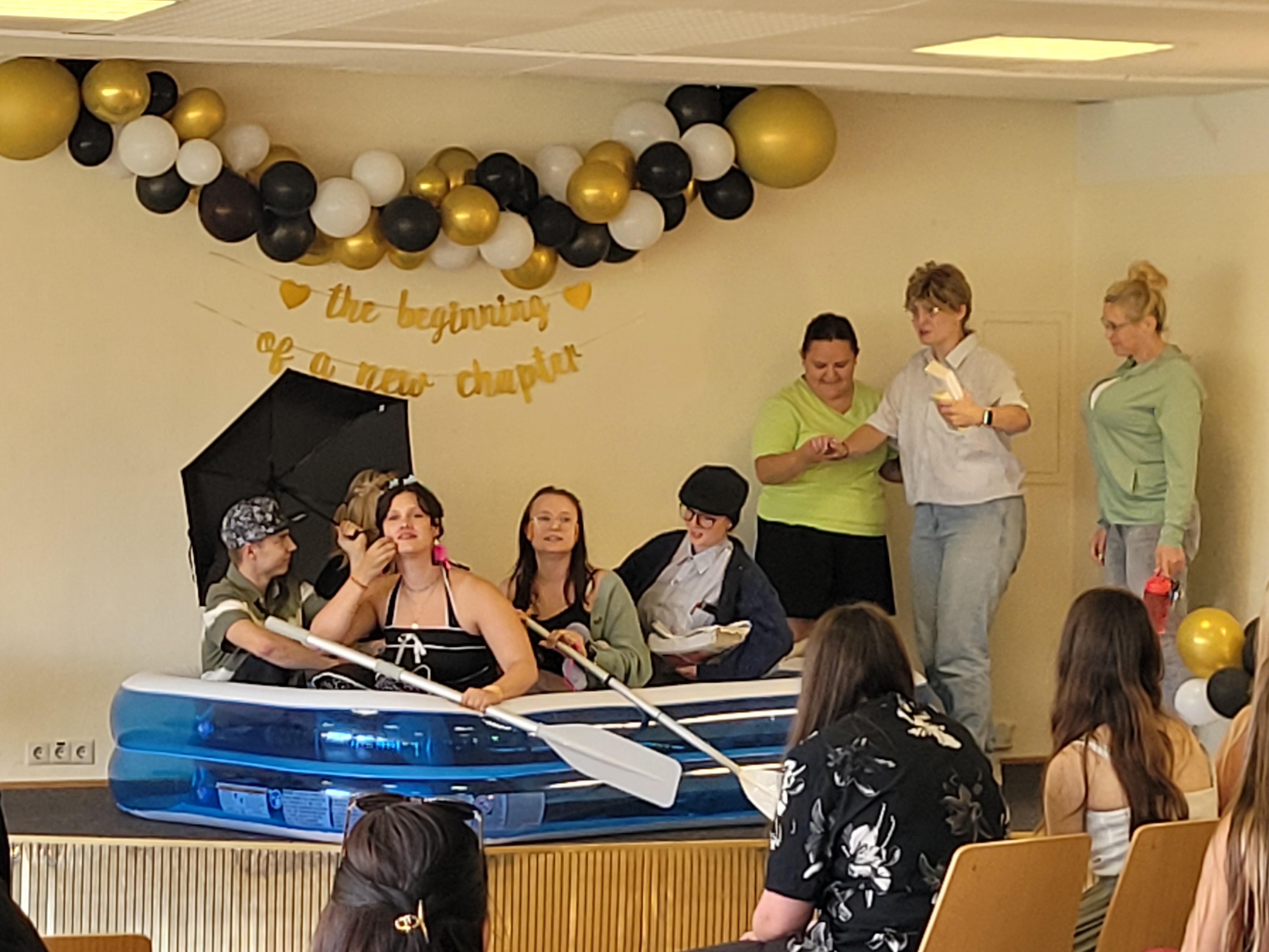 Auf einer Bühne sitzen fünf Personen in einem blauen Schlauchboot, drei weitere Personen stehen daneben. Im Hintergrund ist eine Girlande aus schwarzen, weißen und goldenen Luftballons über dem Spruch "The beginning of a new chapter". Im Vordergrund sitzen Zuschauer auf Stühlen.