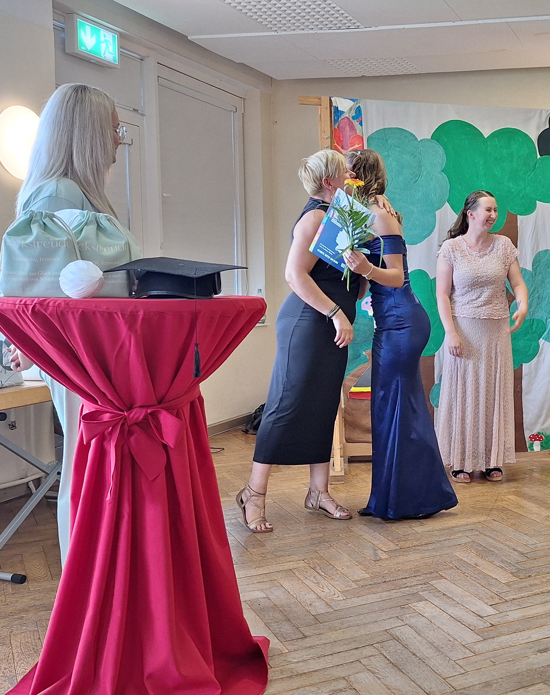 Links steht ein Stehtisch mit einem roten Überzug, auf dem ein Absolventenhut liegt; dahinter steht eine Frau, die zu zwei Frauen in Abendkleidern schaut, die sich umarmen. Die umarmte Frau trägt eine Blume und ein Zeugnis. Rechts steht vor einem Wandbild mit Bäumen eine weitere junge Frau im Abendkleid.