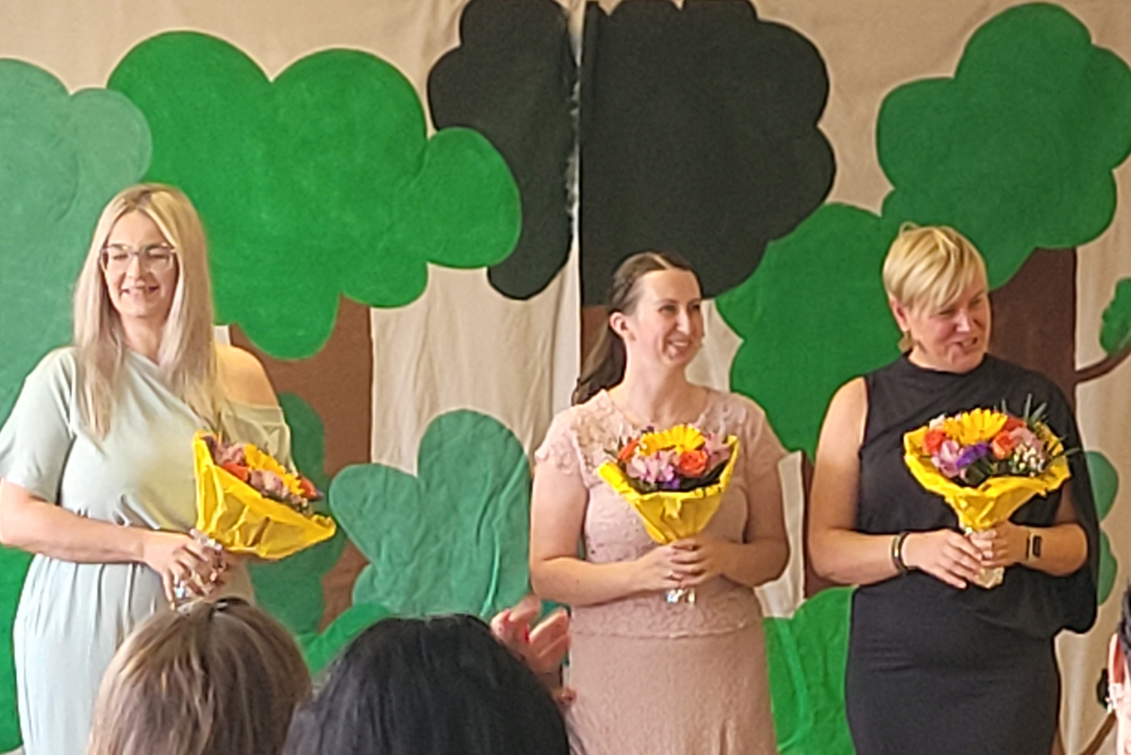 Vor einer mit Bäumen bemalten Wand stehen drei lachende Frauen in Abendkleidern, die alle einen Blumenstrauß in gelbem Papier halten.