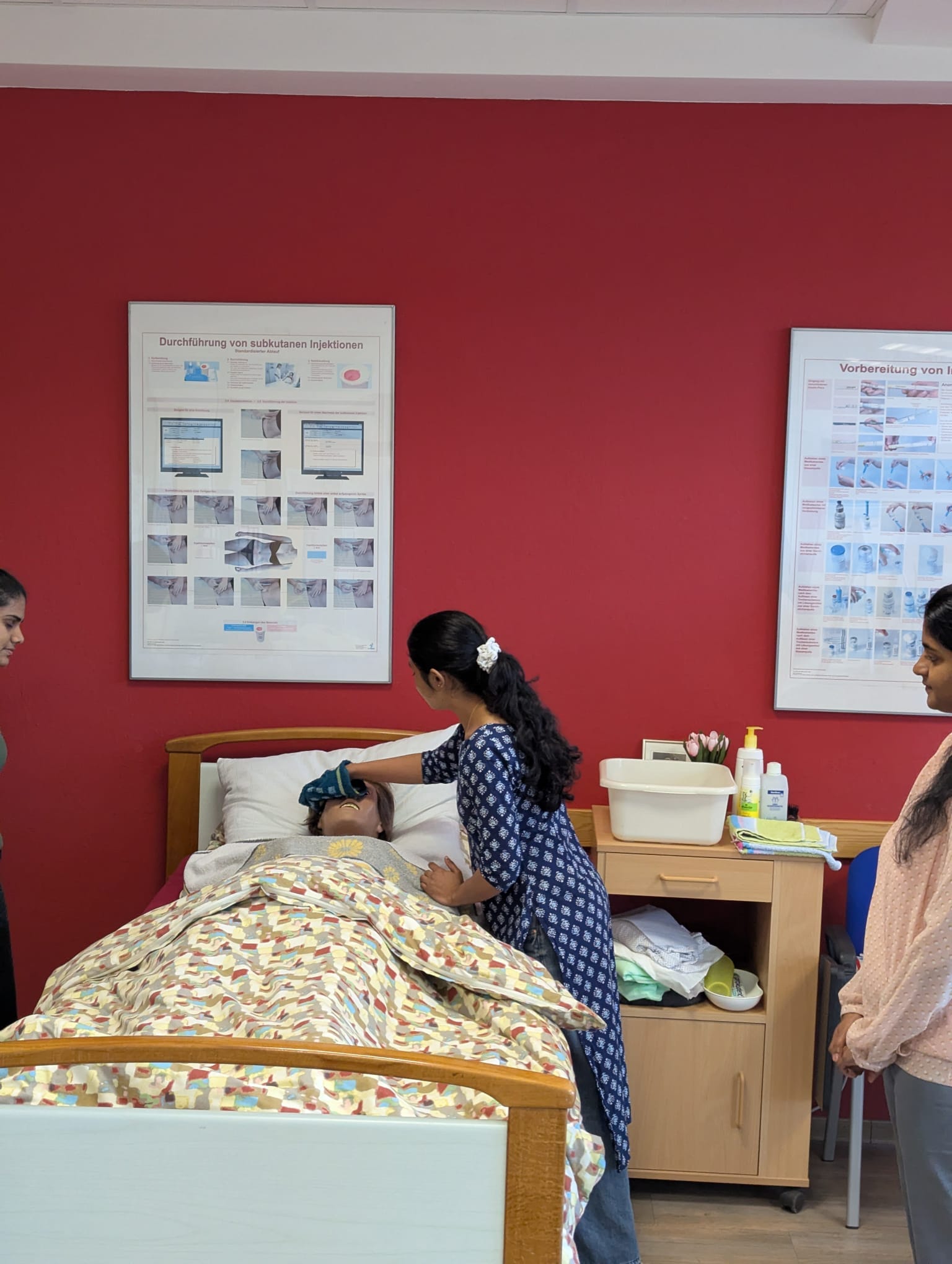 Eine Schülerin wäscht einer Pflegepuppe, die im Bett liegt, mit einem Waschlappen das Gesicht. Zwei Mitschülerinnen schauen ihr dabei über die Schulter. Auf dem Nachttisch steht eine weiße Wanne mit Wasser sowie verschiedene Pflegeutensilien. An der roten Wand hängt ein großes Poster mit dem Titel "Durchführung von subkutanen Injektionen".