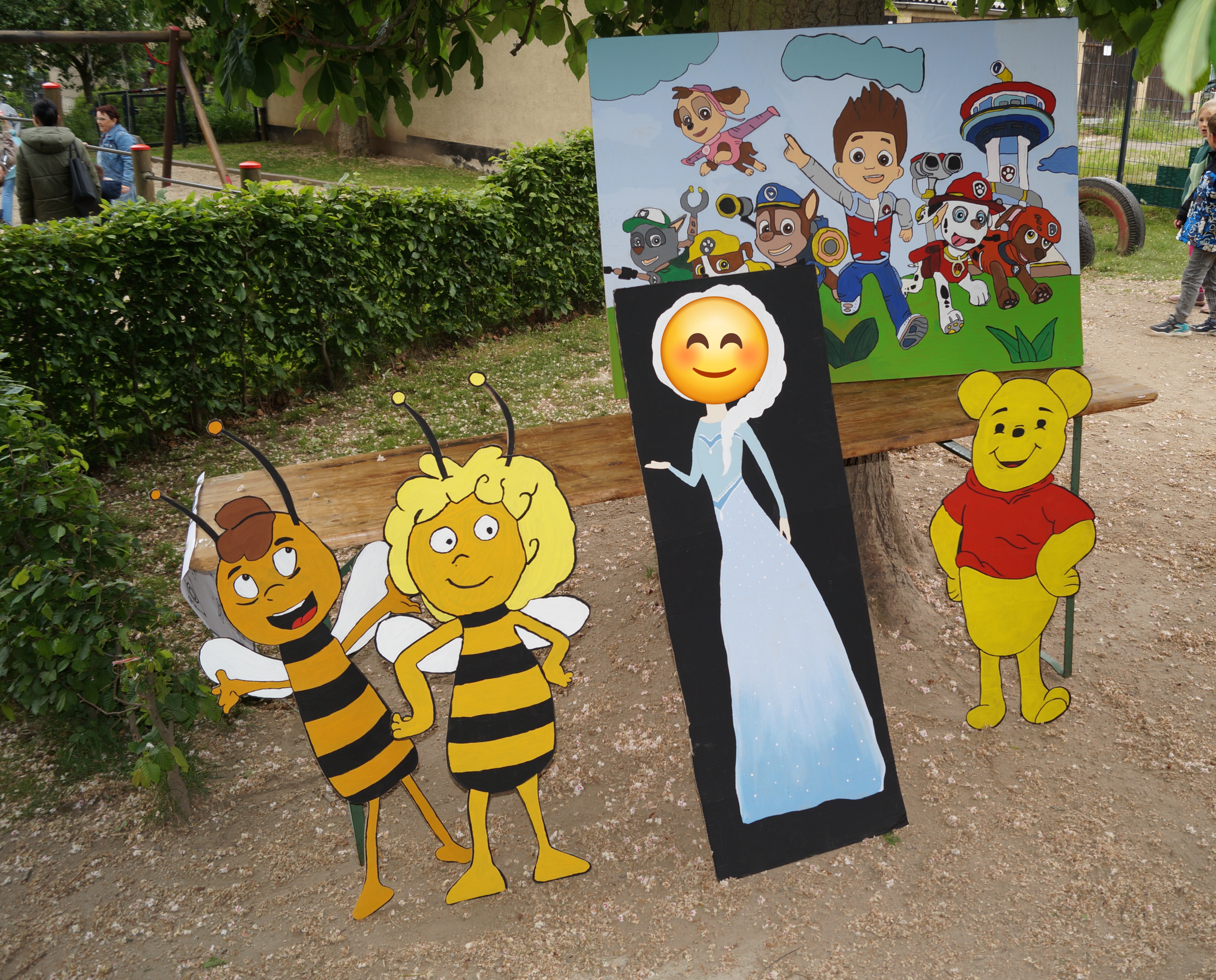 60 Jahre Kita Flax und Krümel – ein Fest, mit allem Drum und Dran Ein Foto zeigt einen spielerischen Aufbau mit Holzfiguren von Biene Maja, Winnie Puuh und einem bemalten Brett mit einer Prinzessin-Figur (mit Emoji-überdecktem Gesicht). Im Hintergrund steht ein weiteres großes Bild mit Figuren der Zeichentrickserie Paw Patrol. Die Szene findet auf einem Außengelände mit Bäumen statt.