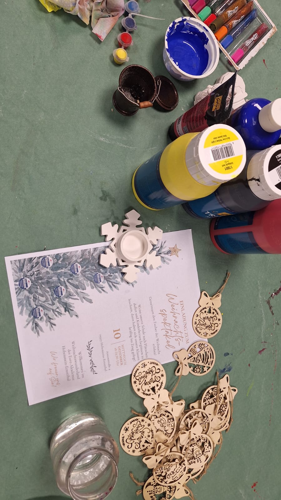 Auf einem grünen Basteltisch liegen bunte Acrylfarben, Pinsel, Wachsmalstifte, eine Einladung zum Weihnachtsspektakel sowie zahlreiche filigrane Holzanhänger in Weihnachtsmotiven und ein weißer Schneeflocken-Teelichthalter.