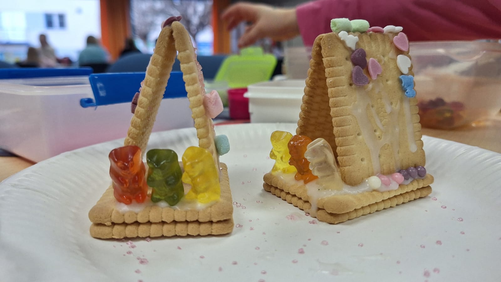 Auf einem Pappteller stehen zwei aus Butterkeksen gebaute Mini-Häuschen, die mit Gummibärchen, Zuckerkugeln und bunten Herzstreuseln verziert sind; im unscharfen Hintergrund sitzen Kinder am Basteltisch.