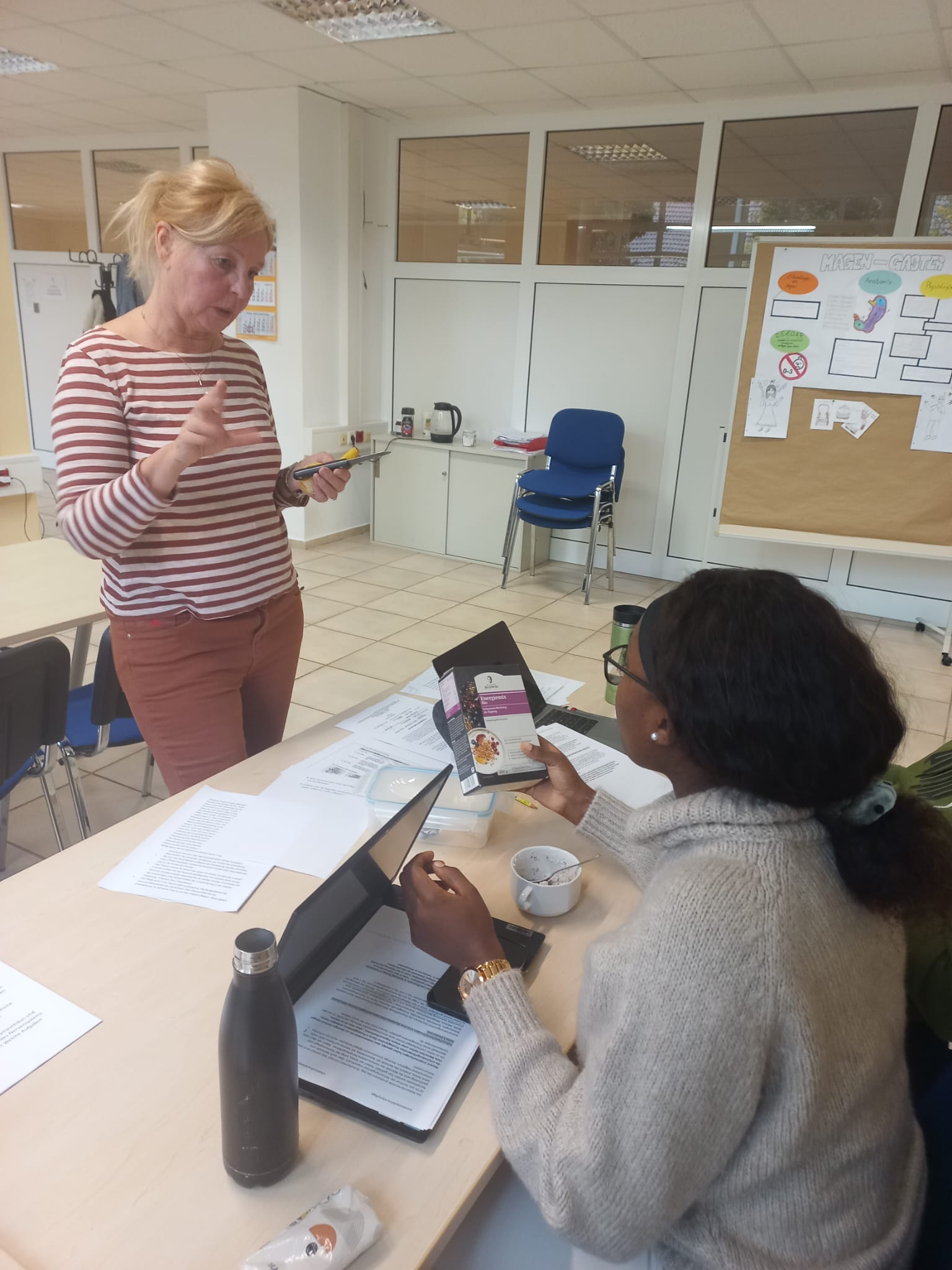 Gesundes Frühstück für Pflegefachmänner*frauen an der Euro Akademie Jena Eine dunkelhäutige Frau mit schwarzen Haaren sitzt an einem Holztisch mit einem Notebook, davor steht eine blonde ältere Frau, die mit ihr redet.