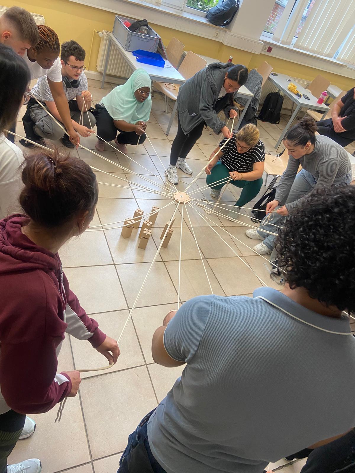 Teambuilding-Übung: Lernende hocken im Kreis und steuern mit langen Schnüren gemeinsam eine Vorrichtung, um Holzklötze zu bewegen.