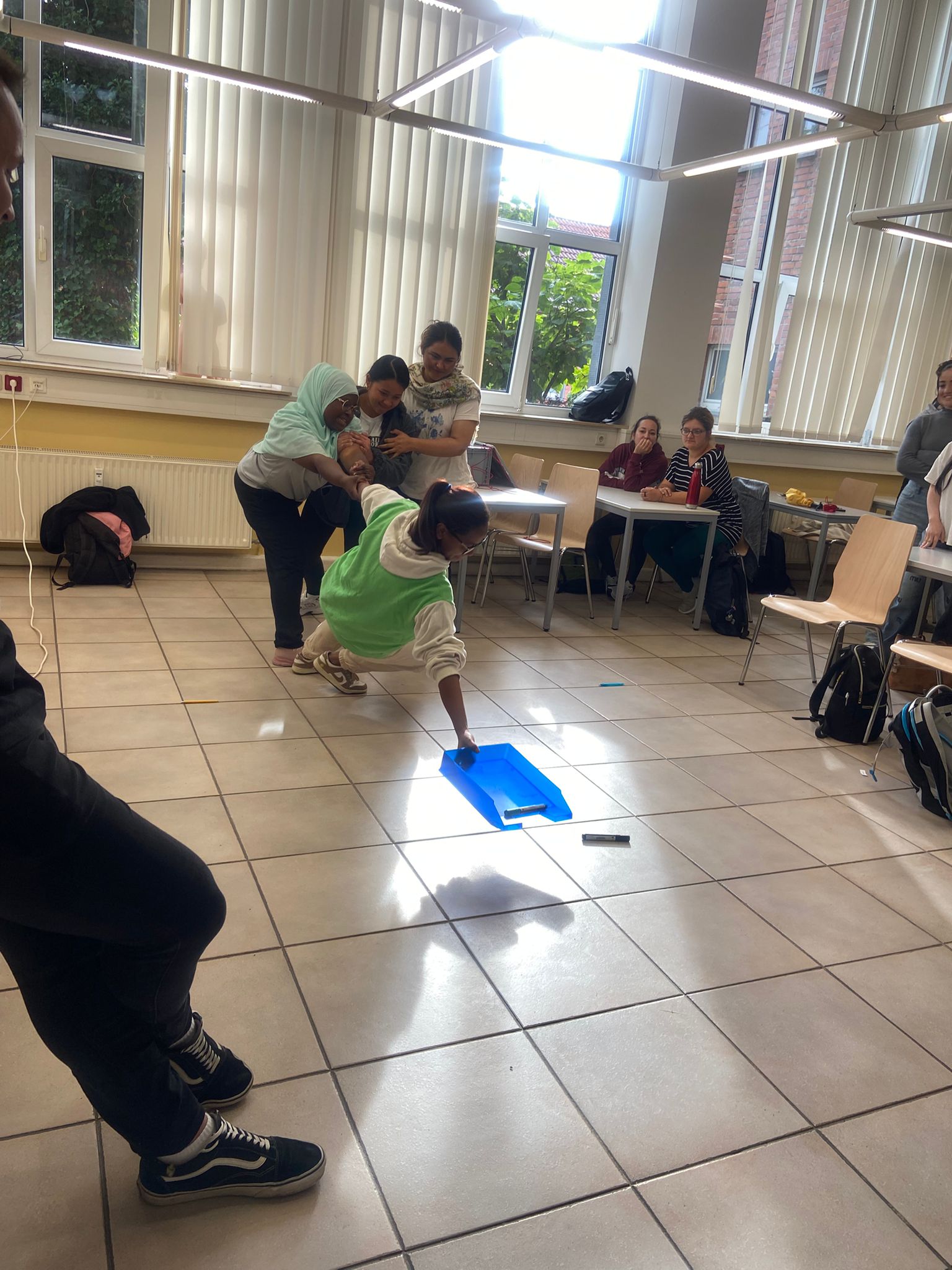 Kooperationsspiel im Klassenraum: mehrere Personen sichern eine Mitspielerin, die sich nach einem blauen Tablett am Boden streckt.