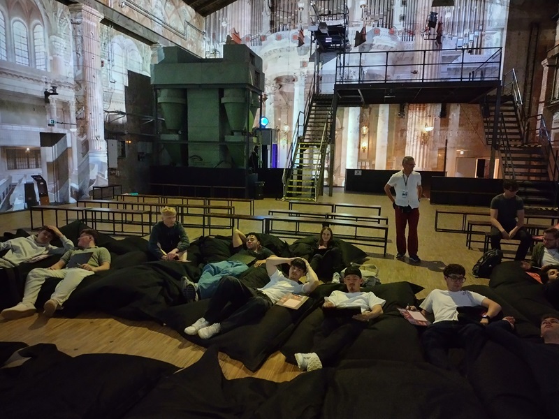 Mehrere Schülerinnen und Schüler liegen entspannt auf großen, dunklen Sitzsäcken in einer geräumigen Halle mit hoher Decke. Im Hintergrund sind industrielle Maschinen, eine Metalltreppe und eine Empore zu sehen. Die Wände sind mit Lichtprojektionen versehen, die architektonische Details und Fenster andeuten. Eine Person steht aufrecht und blickt in Richtung der Kamera.