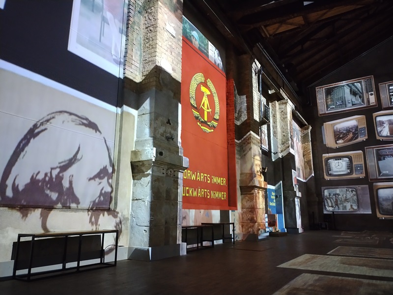 Große rote Fahne mit Hammer, Zirkel und Ährenkranz hängt an einer Backsteinwand in einer Ausstellungshalle. Links davon sind Projektionen historischer Motive und Porträts auf die Wandflächen geworfen. Rechts stehen mehrere alte Fernsehgeräte in Regalen, deren Bildschirme verschiedene Schwarz-weiß-Aufnahmen zeigen. Die Halle hat hohe Decken mit sichtbaren Holzbalken und ist spärlich beleuchtet.