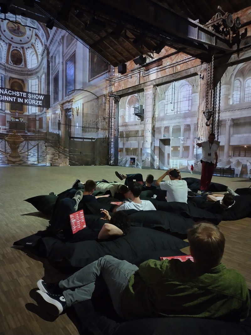 Mehrere Berufsschüler*innen liegen entspannt auf dunklen Sitzsäcken in einem abgedunkelten Raum. Die Wände sind großflächig mit Projektionen historischer Architektur und Kirchenfenstern beleuchtet. Eine Person steht vorne und scheint zu sprechen oder zu präsentieren. Die Atmosphäre wirkt ruhig und konzentriert.