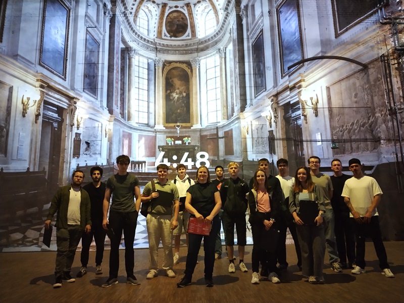 Die Gruppe der Berufsschüler*innen steht in einem Innenraum vor einer großformatigen Wandprojektion, die eine prunkvolle, klassizistische Kirche mit hohen Rundbogenfenstern, Kuppel und Gemälden zeigt und posiert für ein Gruppenbild. Die Personen sind frontal zur Kamera ausgerichtet und lächeln überwiegend. In der Mitte der Projektion ist eine digitale Uhr mit der Zeit '4:48' zu sehen. Die Beleuchtung ist warm und hebt die Gruppe hervor.