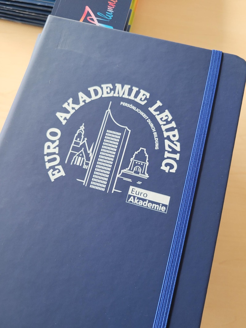 Dunkelblaues Notizbuch mit elastischem Verschlussband. Auf dem Einband ist in weißer Farbe ein kreisförmiger Schriftzug mit der Aufschrift 'EURO AKADEMIE LEIPZIG' sowie eine stilisierte Skyline mit zwei Gebäuden und einem Hochhaus abgebildet. Darunter befindet sich ein kleines, rechteckiges Logo mit der Aufschrift 'Euro Akademie'. Im Hintergrund sind weitere Notizbücher zu erkennen.
