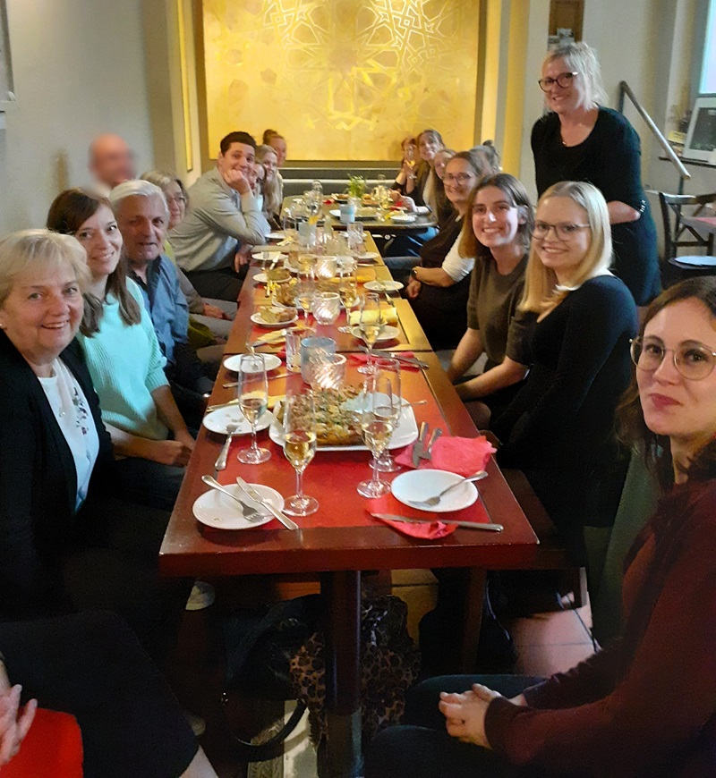 Lange Tafel mit mehreren Absolventinnen und Lehrkräften, die an beiden Seiten sitzen. Auf dem Tisch stehen zahlreiche Gläser mit Getränken, weiße Teller, Besteck und eine große Platte mit Speisen in der Mitte. Im Hintergrund eine gelb beleuchtete Wand mit geometrischem Muster. Die Personen blicken überwiegend in Richtung Kamera, einige lächeln. Eine Person steht am Kopfende des Tisches.