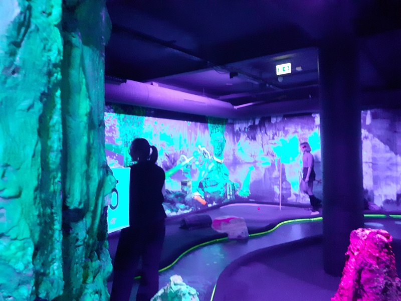 Im Vordergrund stehen mehrere Personen in einem dunklen Raum mit künstlichen Felsen und leuchtenden Linien auf dem Boden. Die Wände sind mit intensiven lilafarbenen und grünen Lichtprojektionen beleuchtet, die abstrakte Muster und Landschaften zeigen. Die Szene wirkt futuristisch und ist von einer starken Farbgebung geprägt.