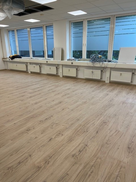 Großer, heller Raum mit frisch verlegtem Holzfußboden, großen Fenstern auf einer Seite und Heizkörpern darunter. Auf der Fensterbank liegen Kabel und Werkzeuge – auch dieser Raum ist im Umbau.