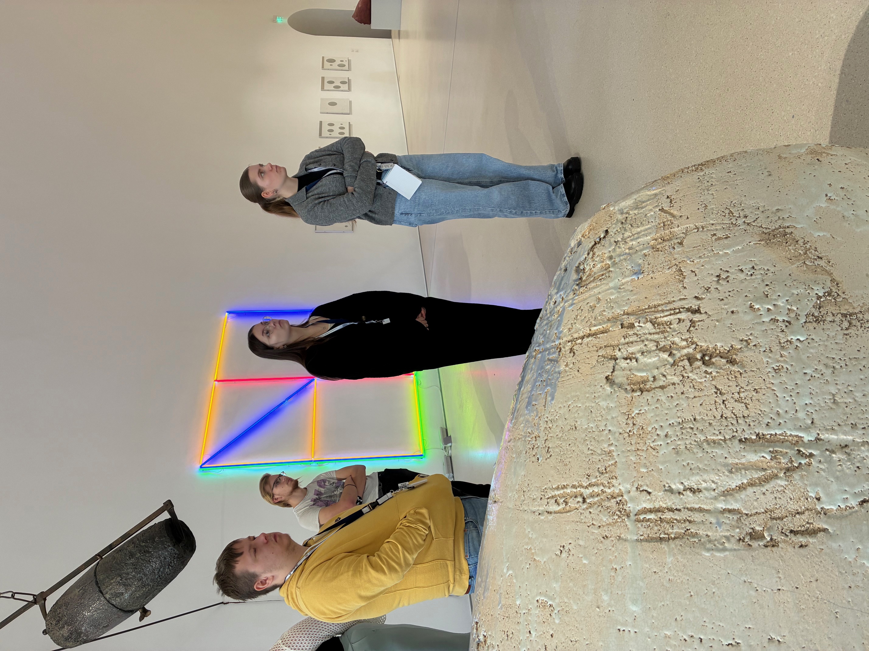 Mehrere Auszubildende zum Erzieher stehen aufmerksam in einem Ausstellungsraum des Kunstmuseums Kloster Unser Lieben Frauen in Magdeburg. Sie betrachten zeitgenössische Kunstwerke im Rahmen einer Exkursion zum Thema frühkindliche ästhetische Bildung. Im Hintergrund leuchten farbige Neonlicht-Installationen in Blau, Rot, Gelb und Grün an einer weißen Museumswand. Rechts sind grafische Arbeiten zu erkennen, links hängt ein großes, dunkles Objekt von der Decke. Im Vordergrund liegt ein helles, strukturreiches Skulpturenelement. Die Lernenden tragen Museumsausweise und wirken konzentriert, während sie die Kunstwerke erkunden. Die Szene vermittelt einen praxisnahen Einblick in kunstpädagogische Bildungsprozesse im Museum.