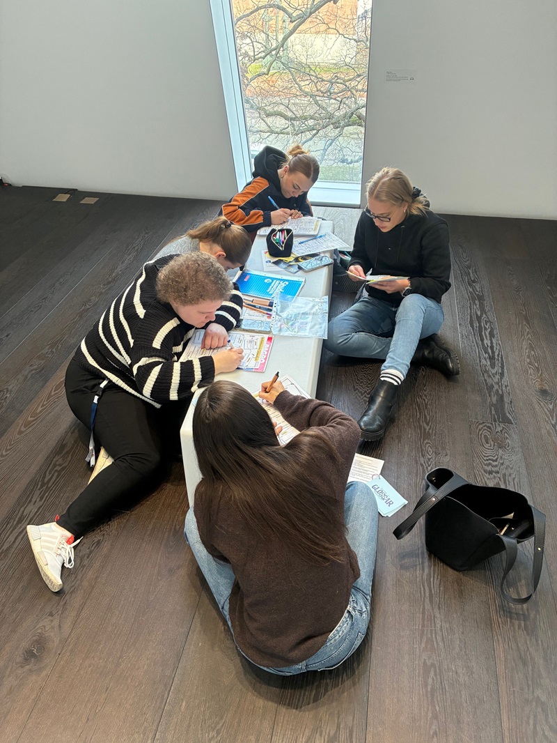 Mehrere Auszubildende zu Erziehern sitzen und liegen auf dem dunklen Holzboden eines hellen Raumes im Kunstmuseum „Kloster Unser Lieben Frauen“ in Magdeburg. Sie sind um einen niedrigen weißen Tisch gruppiert, auf dem verschiedene Arbeitsmaterialien wie Hefte, Stifte und ein schwarzes Cap mit einem bunten Logo liegen. Die Auszubildenden sind in entspannter Haltung vertieft in ihre Arbeit, einige schreiben, andere lesen. Im Hintergrund ist ein großes Fenster mit Blick auf kahle Baumzweige zu sehen, das Tageslicht in den Raum lässt. Neben der Gruppe steht eine schwarze Tasche auf dem Boden. Die Szene vermittelt eine konzentrierte Lernatmosphäre im Rahmen einer Exkursion zum Thema frühkindliche Bildung.