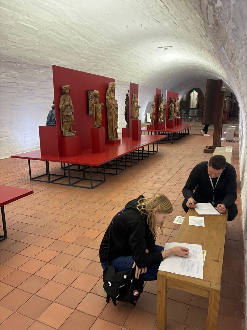 In einem langgestreckten, gewölbten Ausstellungsraum mit weiß verputzten Wänden und terrakottafarbenen Bodenfliesen stehen auf erhöhten, roten Podesten mehrere mittelalterliche Holzskulpturen, die Heilige oder biblische Figuren darstellen. Im Vordergrund sitzen zwei Auszubildende zu Erziehern an einem schlichten Holztisch und bearbeiten schriftliche Unterlagen. Die Atmosphäre wirkt ruhig und konzentriert, passend zu einer Exkursion ins Kunstmuseum „Kloster Unser Lieben Frauen“ in Magdeburg, bei der die Auszubildenden im Rahmen des Unterrichts zum Thema frühkindliche Bildung lernen.