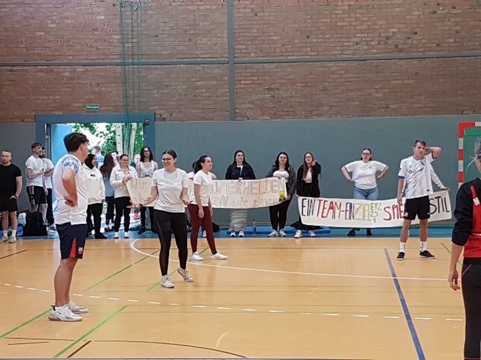 In Tradition: Ein gelungener Start ins neue Schuljahr – Kennenlernen von Schule und Gemeinschaft Am Spielfeldrand stehen die Unterstützer der Teams mit selbst gemalten Transparenten mit motivierenden Worten darauf. Auf dem Volleyballfeld stehen die Spielerinnen und Spieler.