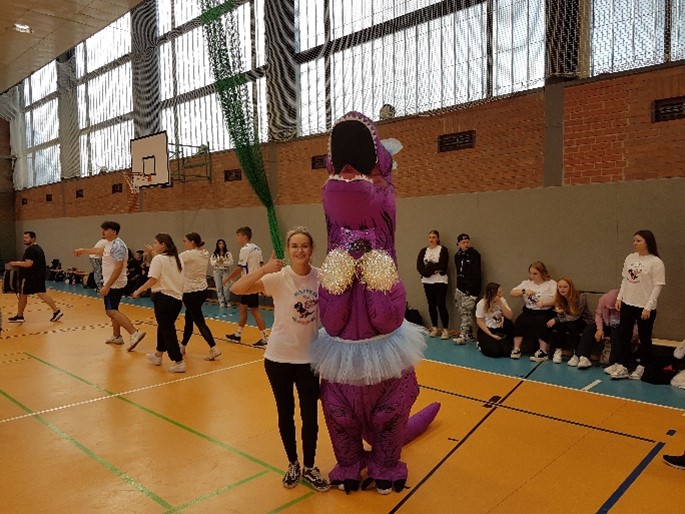 In Tradition: Ein gelungener Start ins neue Schuljahr – Kennenlernen von Schule und Gemeinschaft Ein besonderes Highlight war das lila farbene Dinokostüm der Erzieherklasse 2024, das als Maskottchen das Volleyballturnier begleitete. Neben dem Dino steht eine Auszubildende und zeigt lächelnd einen Daumen nach oben. Im Hintergrund sieht man weitere Teilnehmende in Sportkleidung in der Turnhalle.