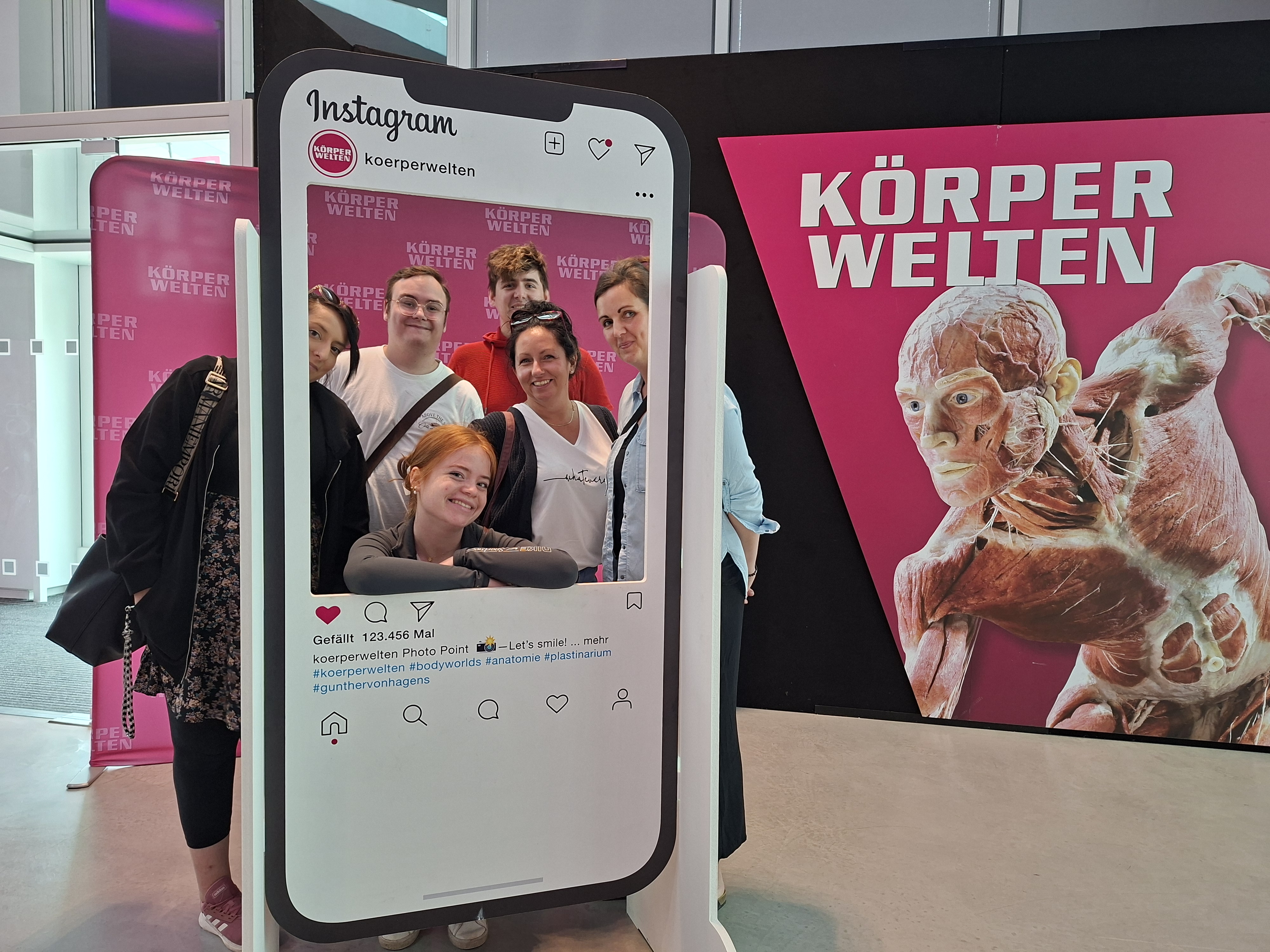 Körperwelten – der Kreislauf des Lebens Eine kleinere Gruppe von Personen posiert fröhlich hinter einem großen Instagram-Fotorahmen in der Ausstellung "Körperwelten". Im Hintergrund ist ein weiteres Plakat mit einer anatomischen Figur sowie der Schriftzug "KÖRPERWELTEN" in Weiß auf pinkem Grund zu sehen.