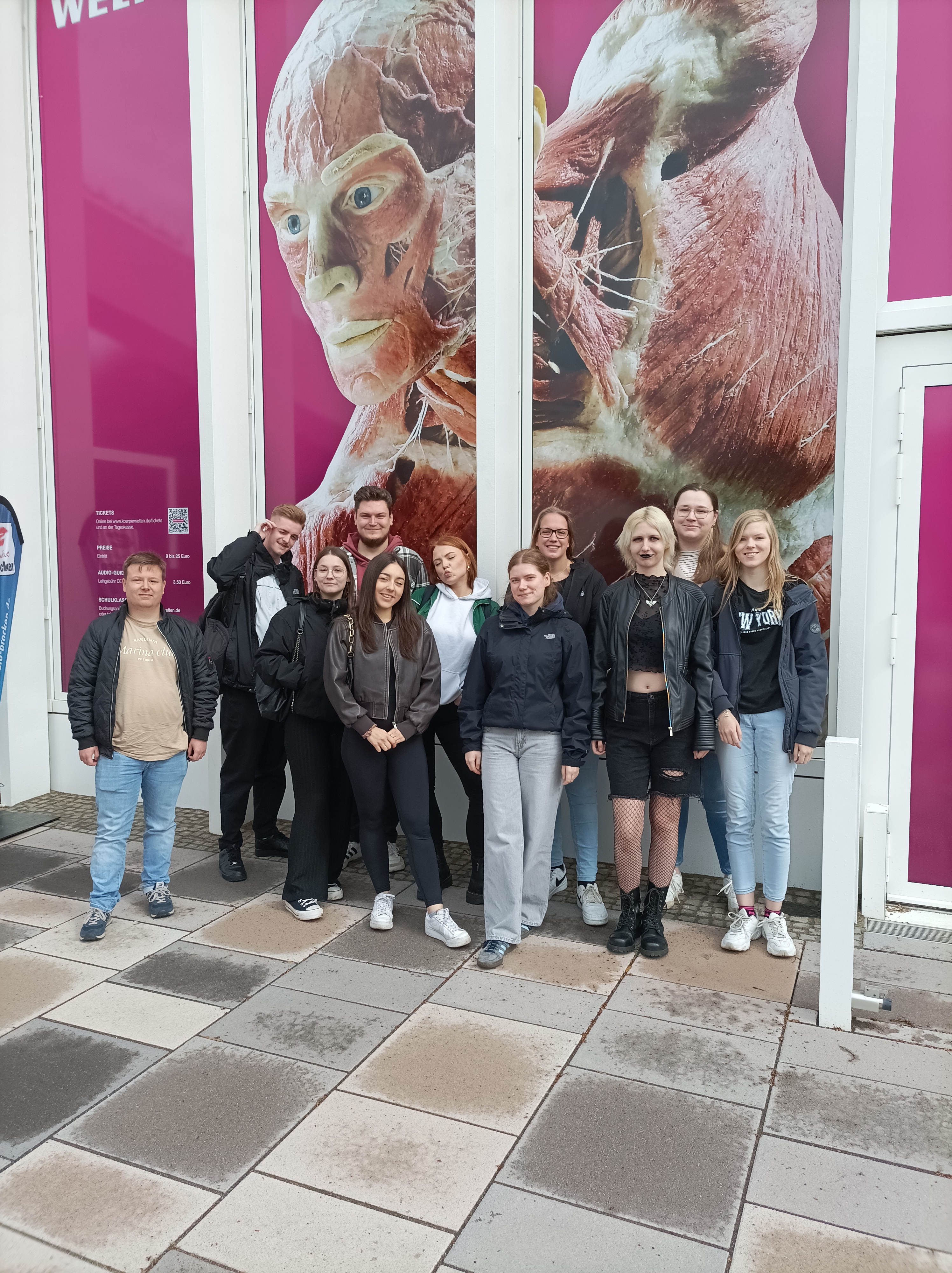 Körperwelten – der Kreislauf des Lebens Eine Gruppe junger Menschen steht lächelnd vor dem Eingang zur Ausstellung "Körperwelten". Hinter ihnen ist ein großes, pinkfarbenes Plakat mit einer plastinierten menschlichen Figur zu sehen. Der Boden ist gepflastert und wirkt leicht feucht.