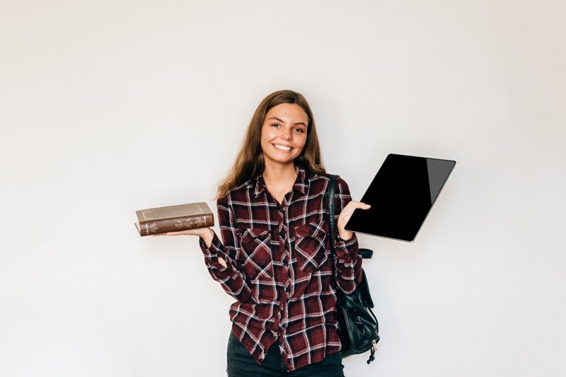 Eine Schülerin hält lächelnd in der linken Hand ein Buch und in der rechten ein Tablet. Sie trägt ein kariertes Holzfällerhemd und hat einen Rucksack um die Schulter geworfen.